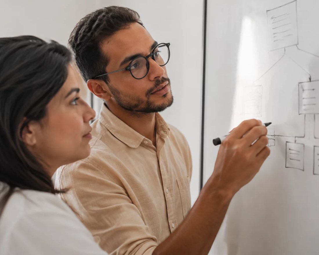 Duas pessoas em pé diante de um quadro branco onde um argumento está desenhado